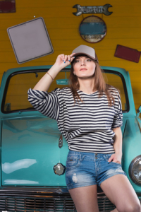 pretty young woman in a cap and a striped shirt and shorts standing near retro blue car in the garage. Holds visor cap