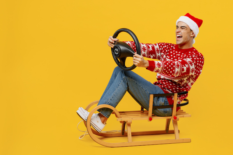 Full body merry excited young man wear red knitted Christmas sweater Santa hat posing sledding hold steering wheel driving car isolated on plain yellow background. Happy New Year 2023 holiday concept