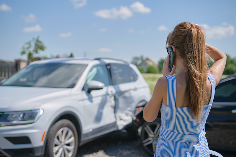 Stressed driver talking on sellphone on roadside near her smashed vehicle calling for emergency service help after car accident. Road safety and insurance concept.