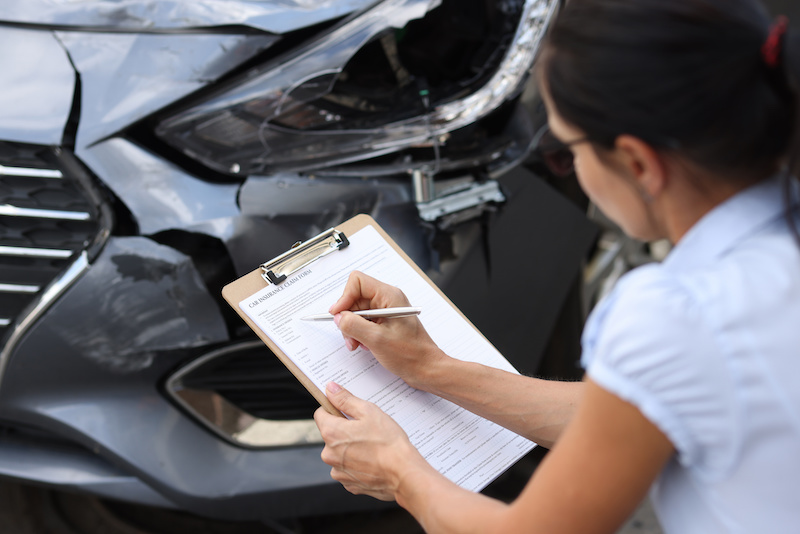 Woman insurance agent filling out paperwork near wrecked car closeup. Estimation of cost of damaged vehicles concept