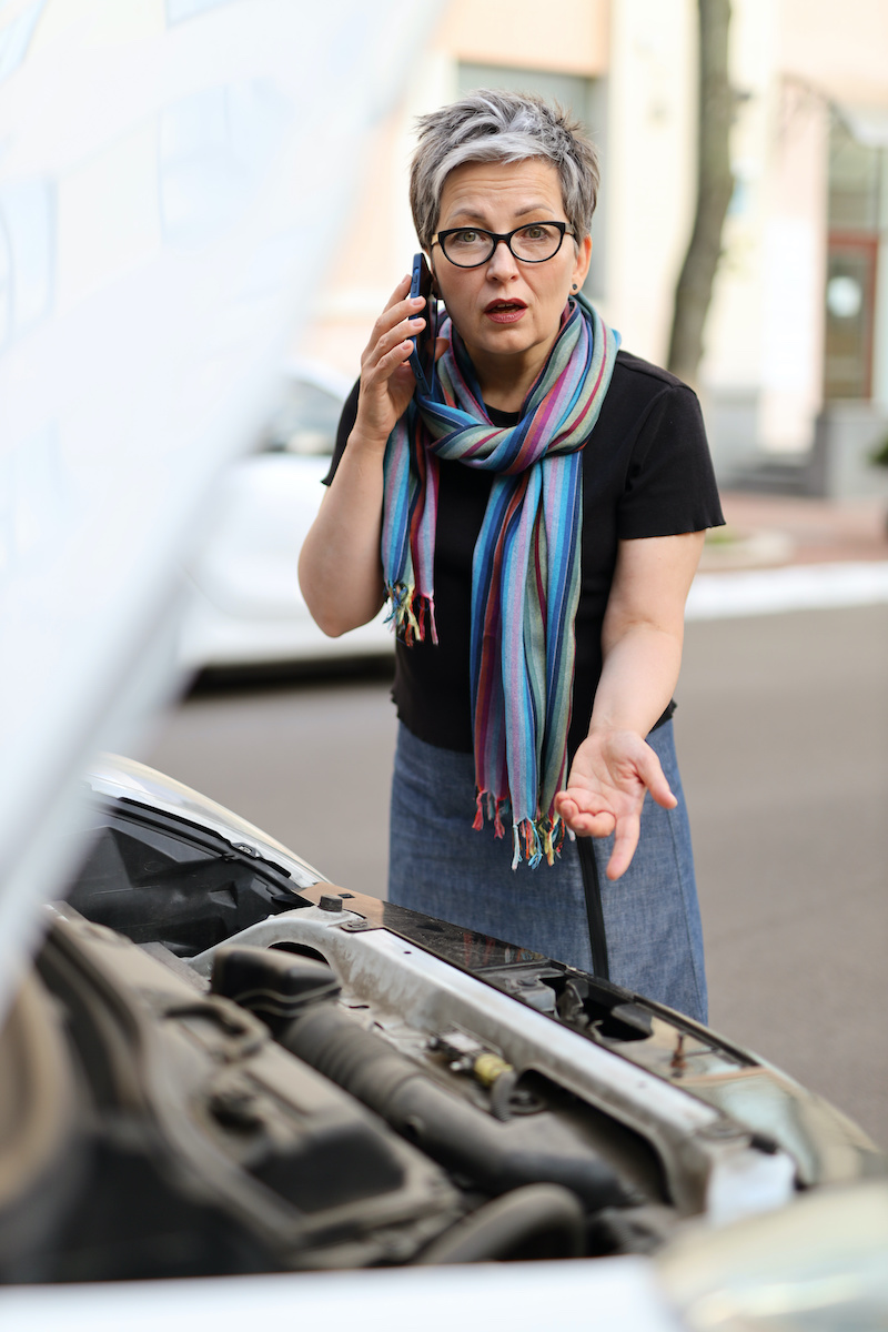 A frustrated senior female driver stands beside her broken-down car, waiting for a mechanic's assistance on the roadside.