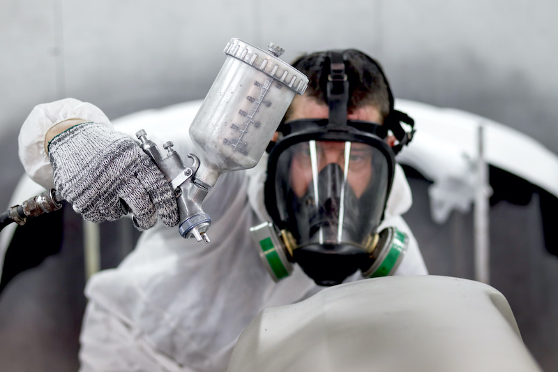 Painting tool in hand of mechanic man painter wearing chemical protective mask while working with auto mechanic painting a car, fixing repairing vehicle at garage automobile repair service shop.