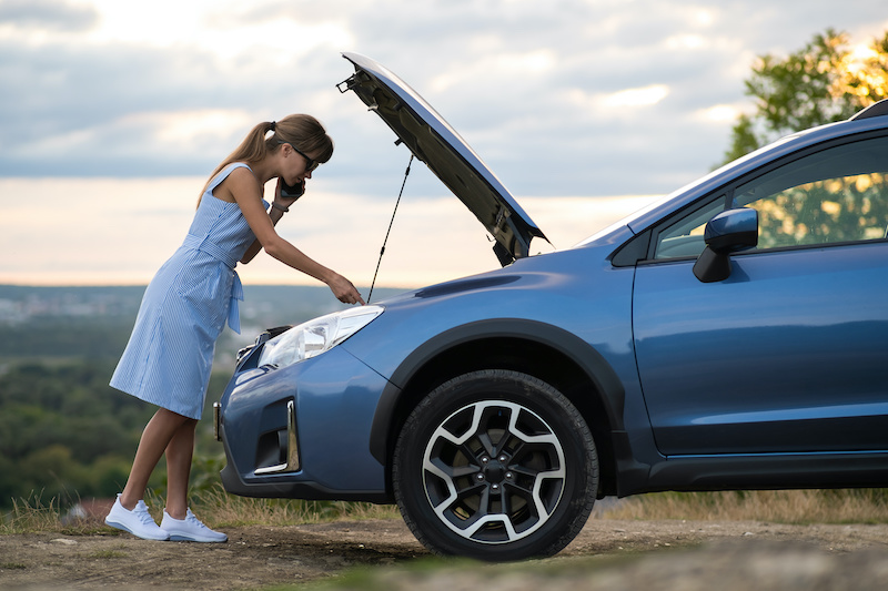 Angry female driver speaking angrily on cell phone with assistance service worker standing near a broken car with popped up hood while inspecting engine having trouble with her vehicle.