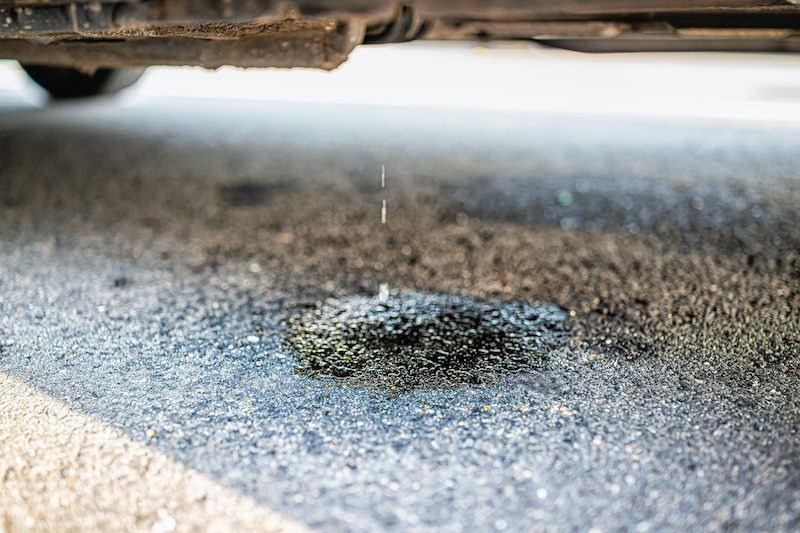 Parked car on street road with macro closeup of vehicle leaking fuel with puddle causing damage to asphalt