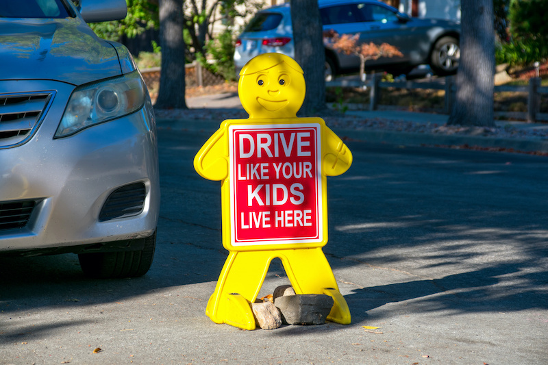 Drive Like Your Kids Live Here road sign in residential neighborhood near parked car. Child like yellow figure with red safety sign on road.