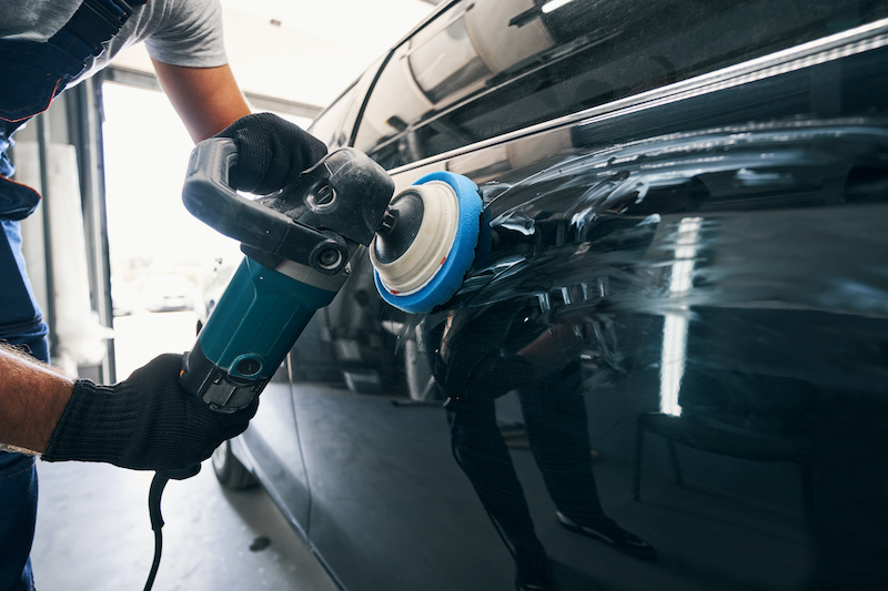 Worker polishing black car side with polisher while holding instrument with both hands in repair service workshop