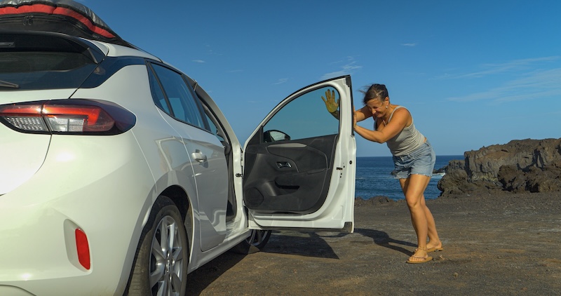 Battling strong coastal winds, a woman tries to close the door of a white car near rocky seashore. Young lady braces against powerful wind, struggling to get the door closed during a stop at the beach