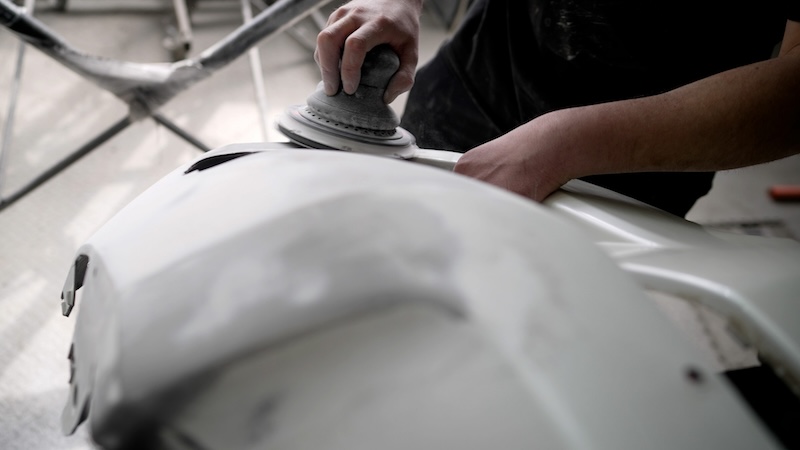 Car mechanic using a pneumatic polishing machine, smoothing a white car bumper in a professional auto repair shop
