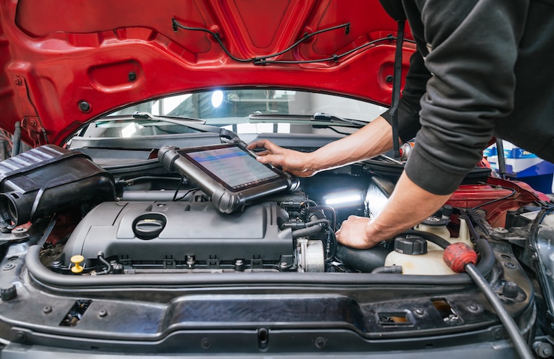 Car mechanic is using a digital tablet to diagnose engine problems on a red car with open hood in a professional auto repair shop, optimizing vehicle performance and ensuring smooth operation