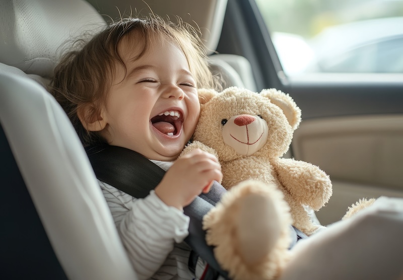 A child sits in the back seat of an SUV, holding a teddy bear and smiling while wearing a white car seat with black trim.