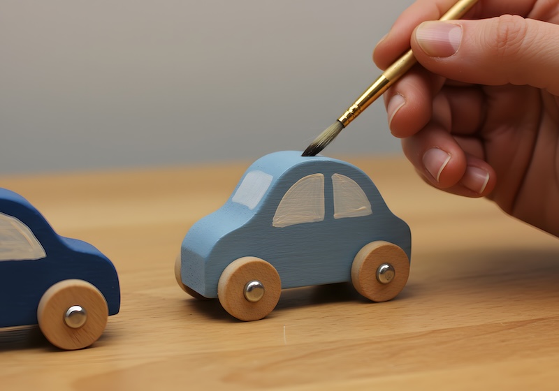 A hand paints a small wooden toy car a light blue color, close-up view, on a light wood surface, likely indoors, daylight, ideal for a hobbyist, craft, or toy-related projects, and commercial or editorial uses.