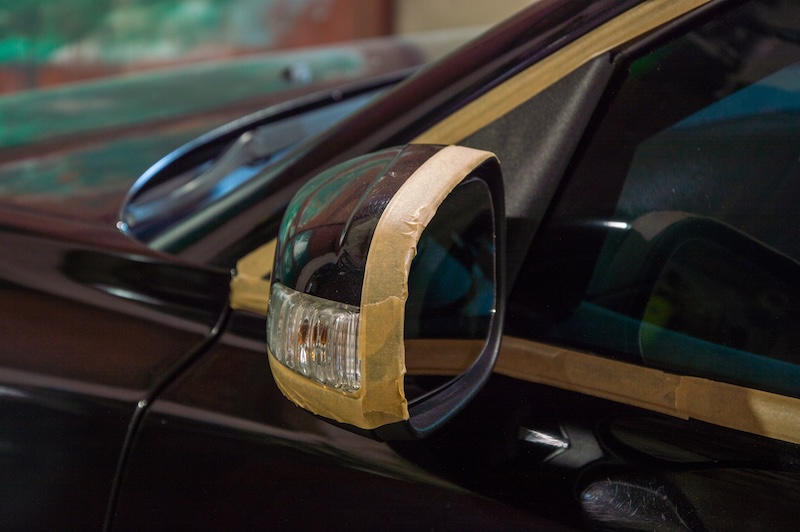 Car polishing is taking place as masking tape is applied carefully around the side mirror, highlighting attention to detail indoors