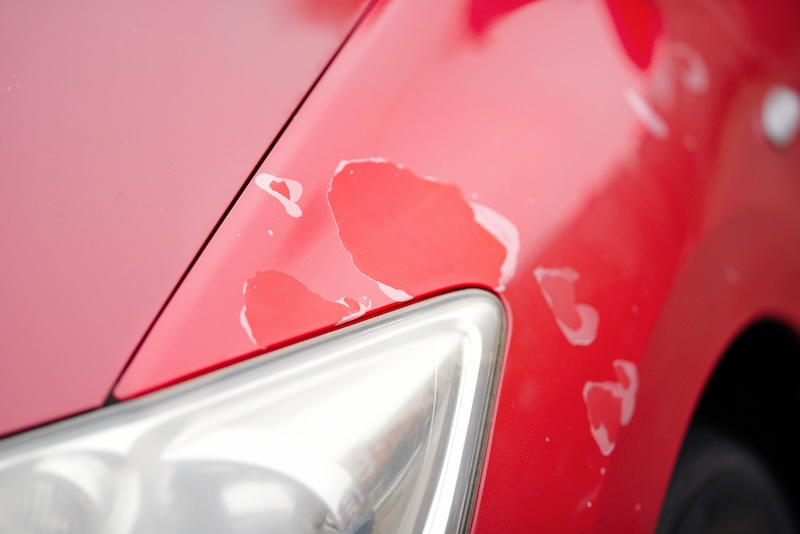 Cracked faded and peeling paint surface exposing clear coat damage and flaking areas on red automobile exterior. Car paint and clear coat damage is primarily caused by UV radiation from the sun