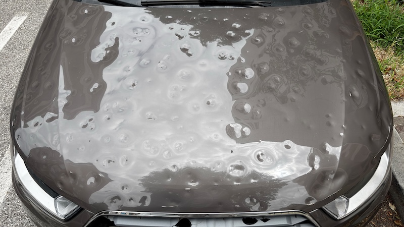 Car hood severely damaged by hail, showing multiple dents and surface imperfections caused by the storm.
