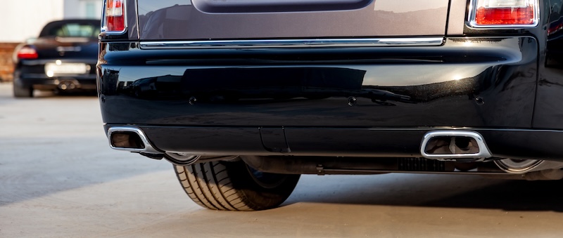 Close-up of a black luxury car bumper of an sedan with two side turbo exhaust pipes outdoors on asphalt. Auto service industry. Air and environment pollution.