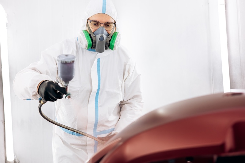 Car painter in protective suit and respirator mask applying new automotive paint to a car body panel in a bright workshop
