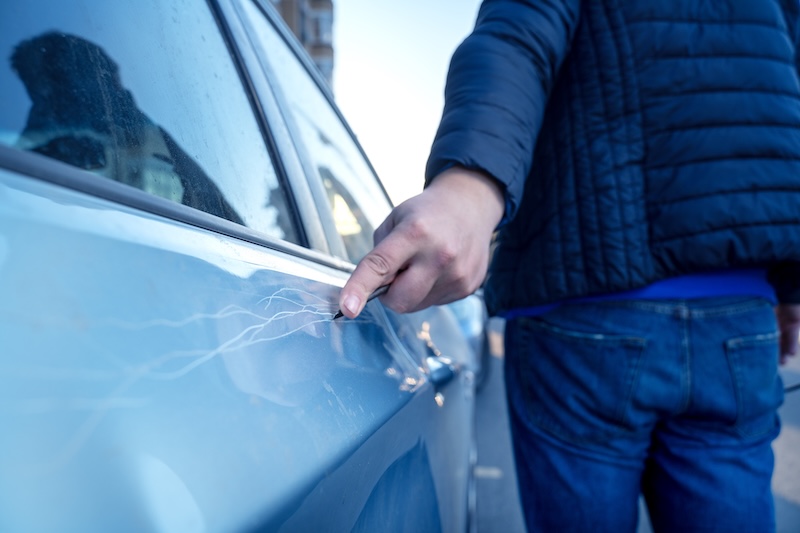 Bad guy scratching the car door with a screwdriver in the parking lot on the street. Damage of property from revenge for treason or betrayal, or threat. Auto insurance fraud or vandalism.