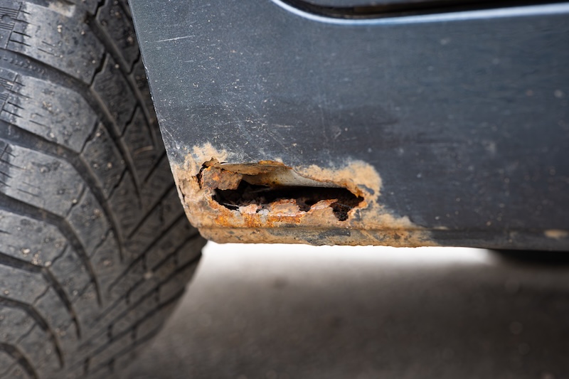 Close up of severe rust damage and corrosion hole on a dark grey car rocker panel near the wheel arch showing decayed metal and peeling paint.