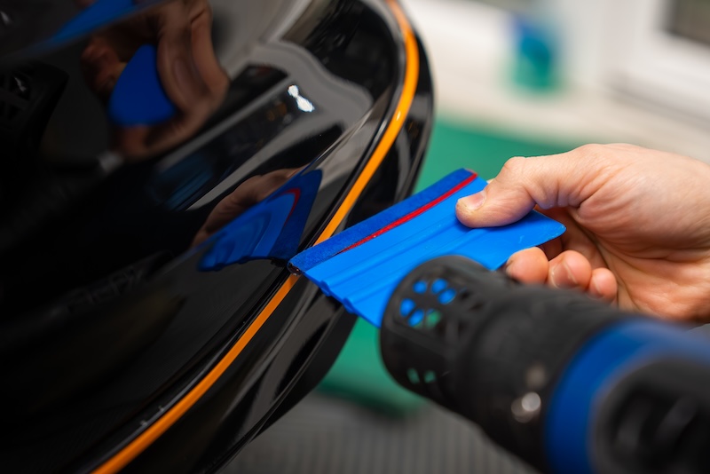 A car technician uses a blue squeegee and a heat gun to fit an orange pinstripe on a glossy black bumper edge in an indoor auto wrap workshop, emphasizing precision.