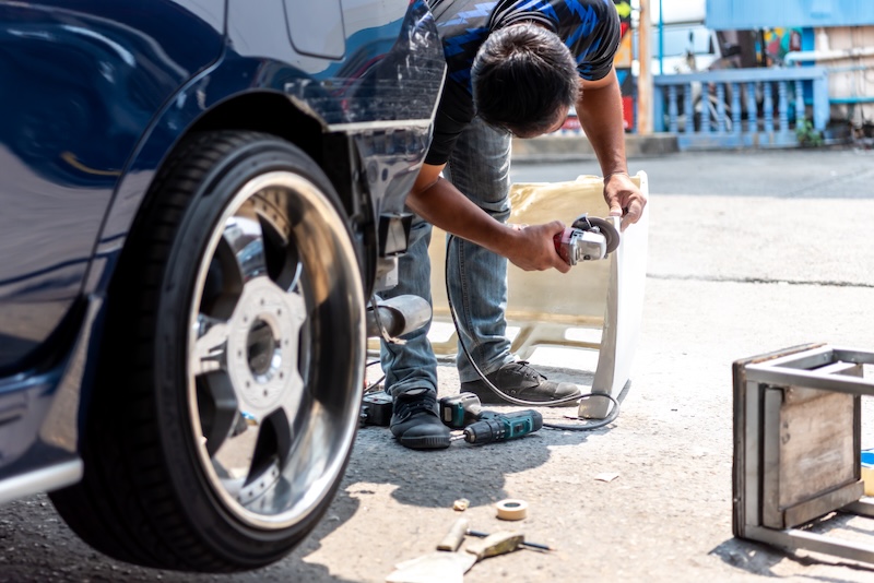 Auto mechanic using angle grinder to adjust bumper during car body repair outdoors with tools and wheel closeup automotive maintenance workshop work