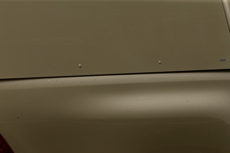 Close-up view of an olive green car trunk and rear panel showing metal surface with subtle reflections, small rivets, panel seams, and faint surface