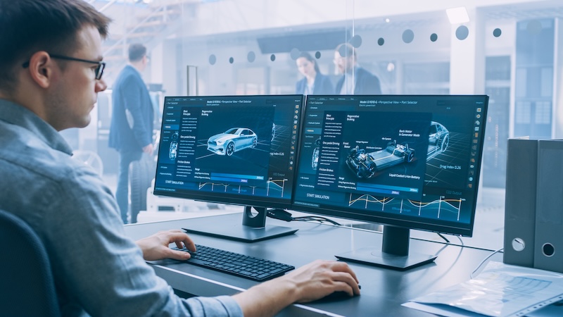 Professional Engineer Works on a Computer with a 3D CAD Software and Tests the Electric Car Chassis Prototype with Wheels, Batteries and Engine Standing in a High Tech Development Laboratory.