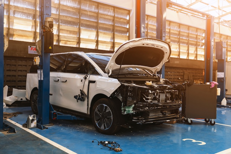 White electric vehicle with open hood and side collision damage on a lift in a professional garage for insurance repair.