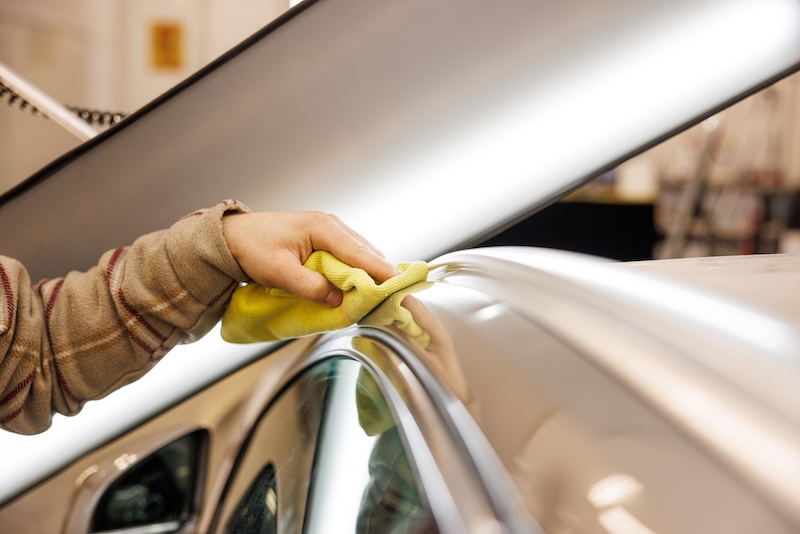 A person is cleaning the automotive exterior of a motor vehicle with a yellow cloth, focusing on the hood, rims, and vehicle doors