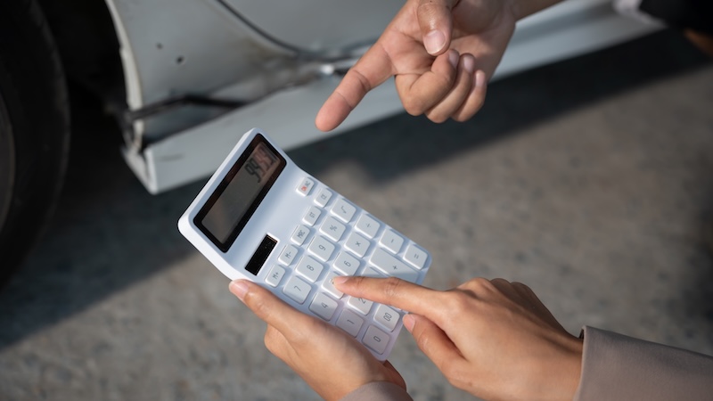 Close-up of hands using calculator while another hand points at a car body, suggesting auto repair cost estimate or car purchase budgeting.