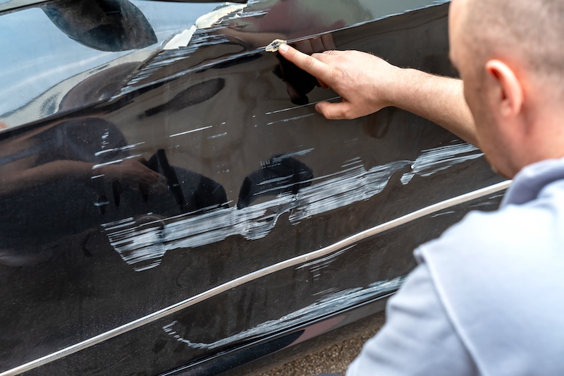 Mechanic kneeling pointing at side damage. Technician examines long scrape and dent along passenger door, gestures to insurance camera, concerned posture, outdoor parking lot, evening light, realistic scene