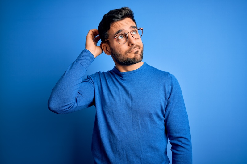 Young handsome man with beard wearing casual sweater and glasses over blue background confuse and wondering about question. Uncertain with doubt, thinking with hand on head. Pensive concept.